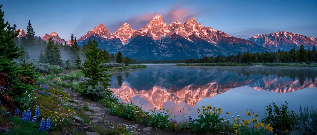 The Grand Teton range reflected in a calm alpine lake at dawn with wildflowers and a hiking trail